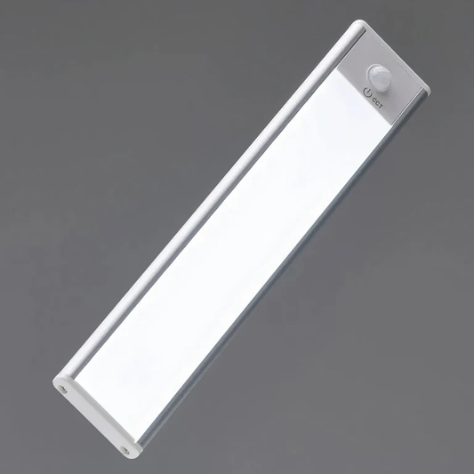 AMBIENTIFY Sora slim rechargeable LED light bar mounted beneath a wooden shelf above a home desk, casting warm even light across the workspace — motion sensor under-cabinet lighting in brushed aluminium.