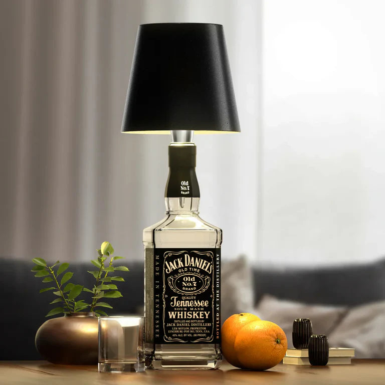 AMBIENTIFY Bottle wireless LED lamp fitted onto a wine bottle, placed on an outdoor garden table surrounded by natural greenery. Soft warm ambient glow, touch-dimmable in 3 levels, USB-C rechargeable table lamp for home décor.