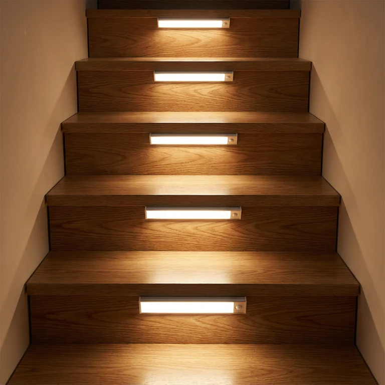 AMBIENTIFY Sora slim rechargeable LED light bar mounted beneath a wooden shelf above a home desk, casting warm even light across the workspace — motion sensor under-cabinet lighting in brushed aluminium.