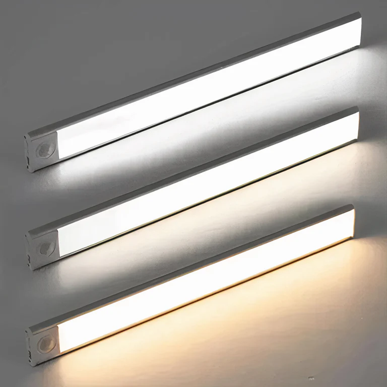 AMBIENTIFY Sora slim rechargeable LED light bar mounted beneath a wooden shelf above a home desk, casting warm even light across the workspace — motion sensor under-cabinet lighting in brushed aluminium.