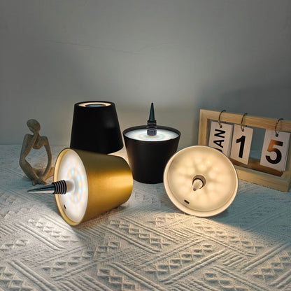 AMBIENTIFY Bottle wireless LED lamp fitted onto a wine bottle, placed on an outdoor garden table surrounded by natural greenery. Soft warm ambient glow, touch-dimmable in 3 levels, USB-C rechargeable table lamp for home décor.