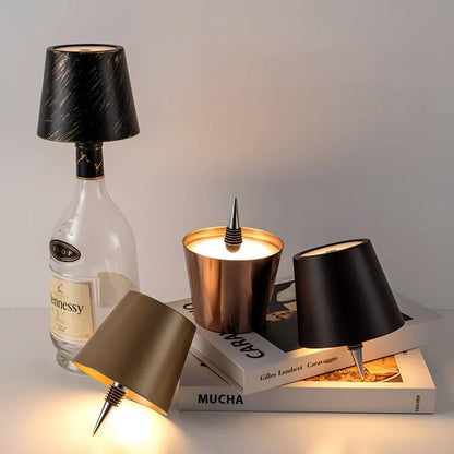 AMBIENTIFY Bottle wireless LED lamp fitted onto a wine bottle, placed on an outdoor garden table surrounded by natural greenery. Soft warm ambient glow, touch-dimmable in 3 levels, USB-C rechargeable table lamp for home décor.