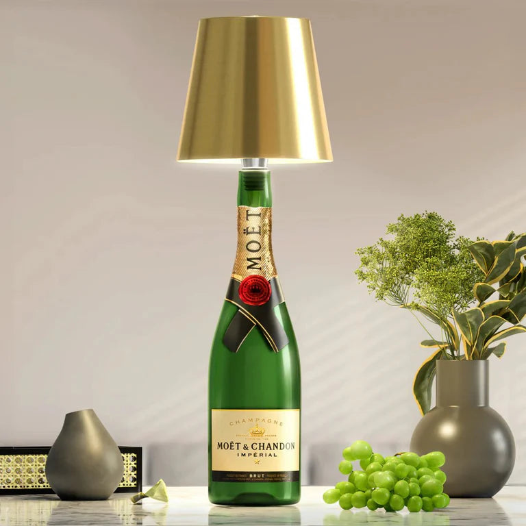 AMBIENTIFY Bottle wireless LED lamp fitted onto a wine bottle, placed on an outdoor garden table surrounded by natural greenery. Soft warm ambient glow, touch-dimmable in 3 levels, USB-C rechargeable table lamp for home décor.