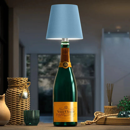 AMBIENTIFY Bottle wireless LED lamp fitted onto a wine bottle, placed on an outdoor garden table surrounded by natural greenery. Soft warm ambient glow, touch-dimmable in 3 levels, USB-C rechargeable table lamp for home décor.