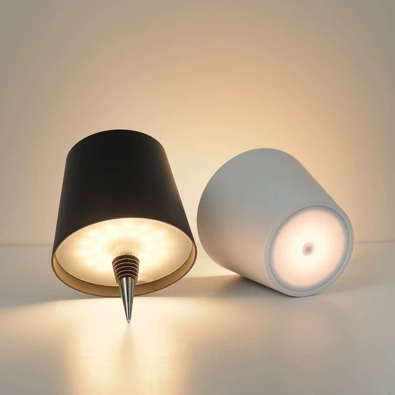 AMBIENTIFY Bottle wireless LED lamp fitted onto a wine bottle, placed on an outdoor garden table surrounded by natural greenery. Soft warm ambient glow, touch-dimmable in 3 levels, USB-C rechargeable table lamp for home décor.