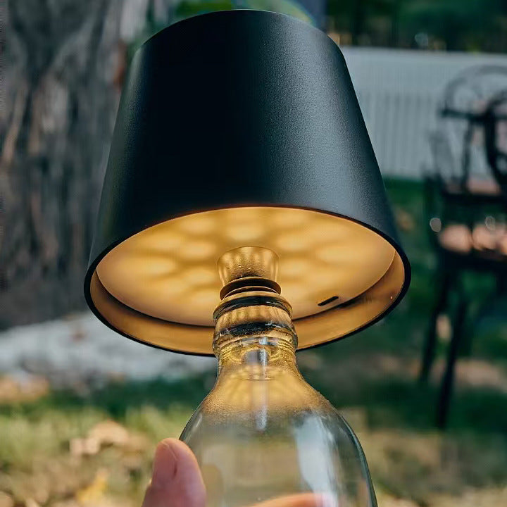 AMBIENTIFY Bottle wireless LED lamp fitted onto a wine bottle, placed on an outdoor garden table surrounded by natural greenery. Soft warm ambient glow, touch-dimmable in 3 levels, USB-C rechargeable table lamp for home décor.