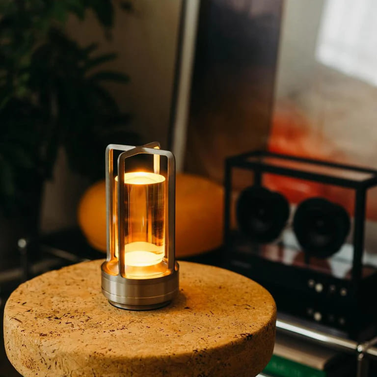 AMBIENTIFY Retro cordless retro table lantern lamp casting a warm amber glow, placed on a wooden tray. Slender aluminium frame with cylindrical acrylic glass shade. Atmospheric home décor lighting for bedroom, living room or outdoor use.