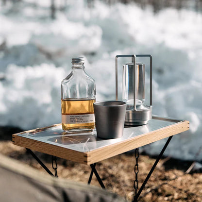 AMBIENTIFY Retro cordless retro table lantern lamp casting a warm amber glow, placed on a wooden tray. Slender aluminium frame with cylindrical acrylic glass shade. Atmospheric home décor lighting for bedroom, living room or outdoor use.