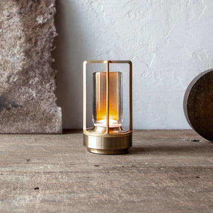 AMBIENTIFY Retro cordless retro table lantern lamp casting a warm amber glow, placed on a wooden tray. Slender aluminium frame with cylindrical acrylic glass shade. Atmospheric home décor lighting for bedroom, living room or outdoor use.