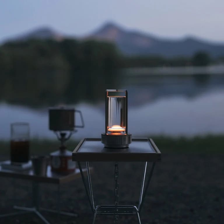 AMBIENTIFY Retro cordless retro table lantern lamp casting a warm amber glow, placed on a wooden tray. Slender aluminium frame with cylindrical acrylic glass shade. Atmospheric home décor lighting for bedroom, living room or outdoor use.