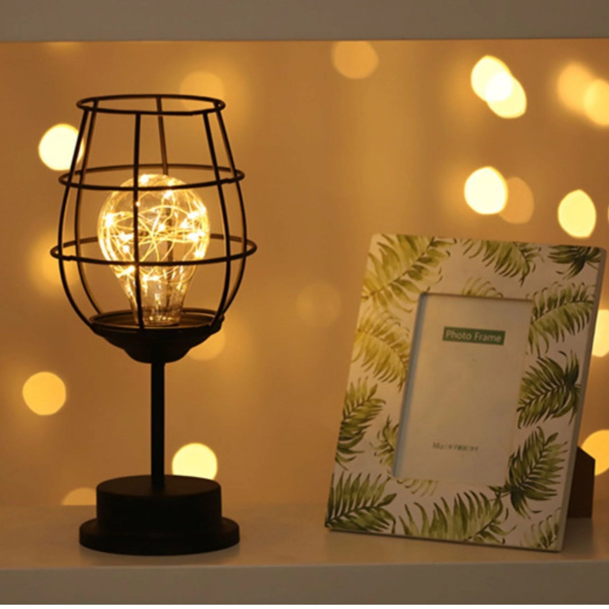 AMBIENTIFY Velora cordless wire-cage table lamp with warm amber LED glow placed on a wooden side table, casting golden light through a sculptural black steel frame — cable-free home accent lighting for any room.