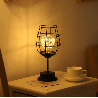 AMBIENTIFY Velora cordless wire-cage table lamp with warm amber LED glow placed on a wooden side table, casting golden light through a sculptural black steel frame — cable-free home accent lighting for any room.