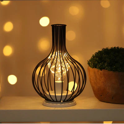 AMBIENTIFY Velora cordless wire-cage table lamp with warm amber LED glow placed on a wooden side table, casting golden light through a sculptural black steel frame — cable-free home accent lighting for any room.