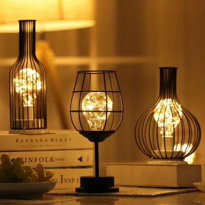 AMBIENTIFY Velora cordless wire-cage table lamp with warm amber LED glow placed on a wooden side table, casting golden light through a sculptural black steel frame — cable-free home accent lighting for any room.