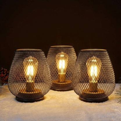 Ambientify Vira cordless metal mesh cage table lamp with a warm Edison-style bulb glowing through a diamond-pattern shade, placed on a wooden surface beside dried flowers and stacked books in a contemporary home interior.