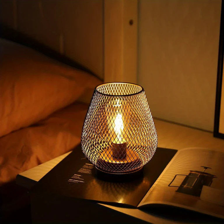 Ambientify Vira cordless metal mesh cage table lamp with a warm Edison-style bulb glowing through a diamond-pattern shade, placed on a wooden surface beside dried flowers and stacked books in a contemporary home interior.