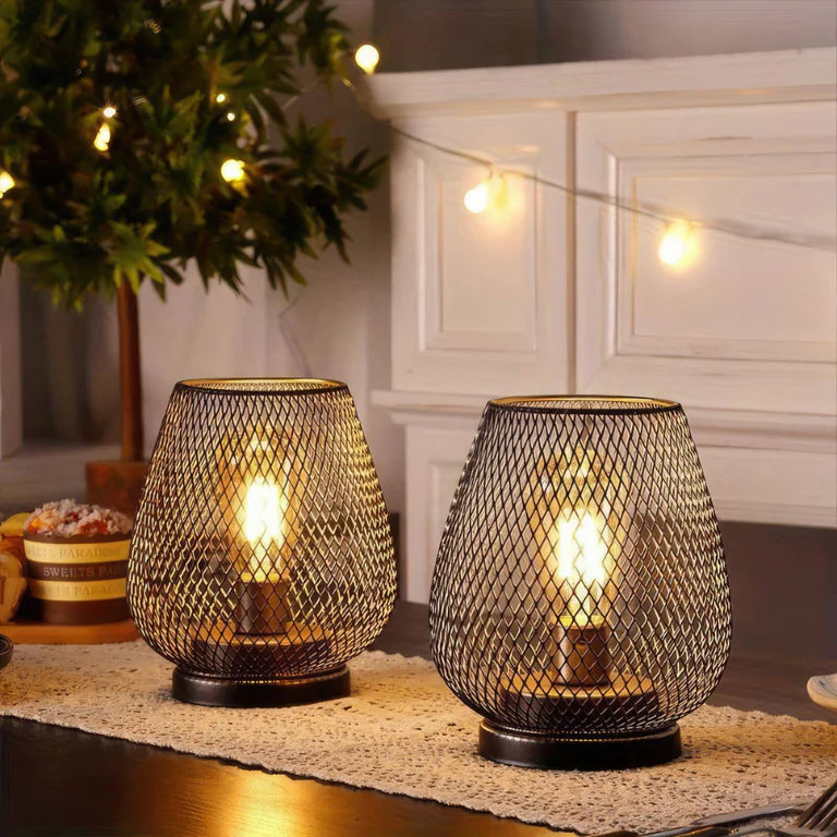 Ambientify Vira cordless metal mesh cage table lamp with a warm Edison-style bulb glowing through a diamond-pattern shade, placed on a wooden surface beside dried flowers and stacked books in a contemporary home interior.