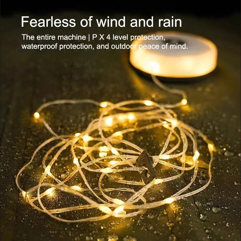 Campify String fairy light reel held in hand at night, showing warm amber LED string lights extended across dark outdoor ground, compact hub glowing with blue charge indicator, IP67 waterproof camping and home fairy lights.