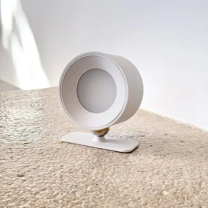 LUNIFY 360 wireless rechargeable LED spotlight with a circular concentric ring surround, mounted on a wall and casting warm amber light in a minimal interior — equally suited as a freestanding spotlight on a shelf or side table.