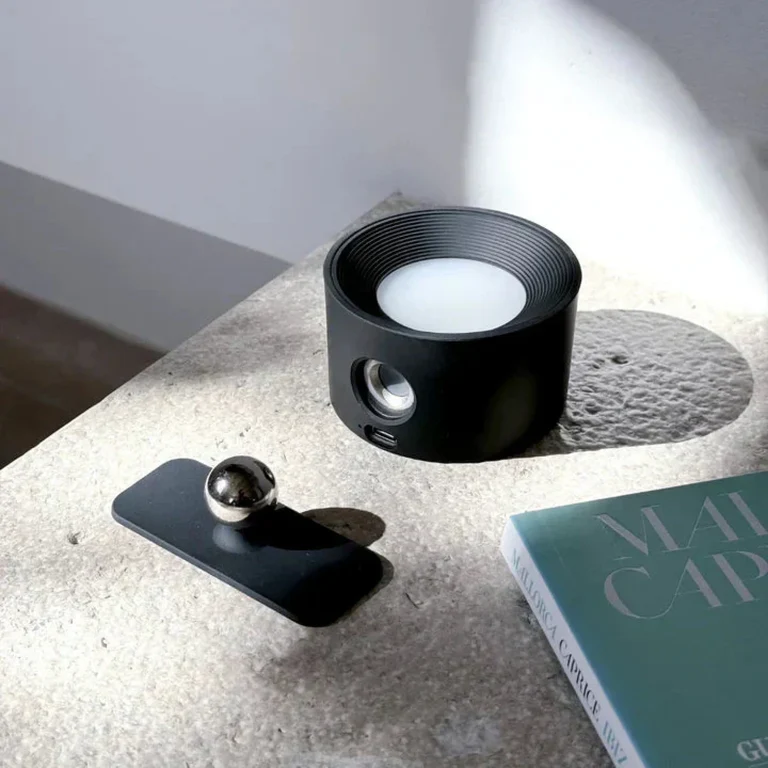 LUNIFY 360 wireless rechargeable LED spotlight with a circular concentric ring surround, mounted on a wall and casting warm amber light in a minimal interior — equally suited as a freestanding spotlight on a shelf or side table.
