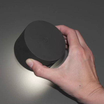 LUNIFY 360 wireless rechargeable LED spotlight with a circular concentric ring surround, mounted on a wall and casting warm amber light in a minimal interior — equally suited as a freestanding spotlight on a shelf or side table.