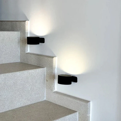 LUNIFY 360 wireless rechargeable LED spotlight with a circular concentric ring surround, mounted on a wall and casting warm amber light in a minimal interior — equally suited as a freestanding spotlight on a shelf or side table.