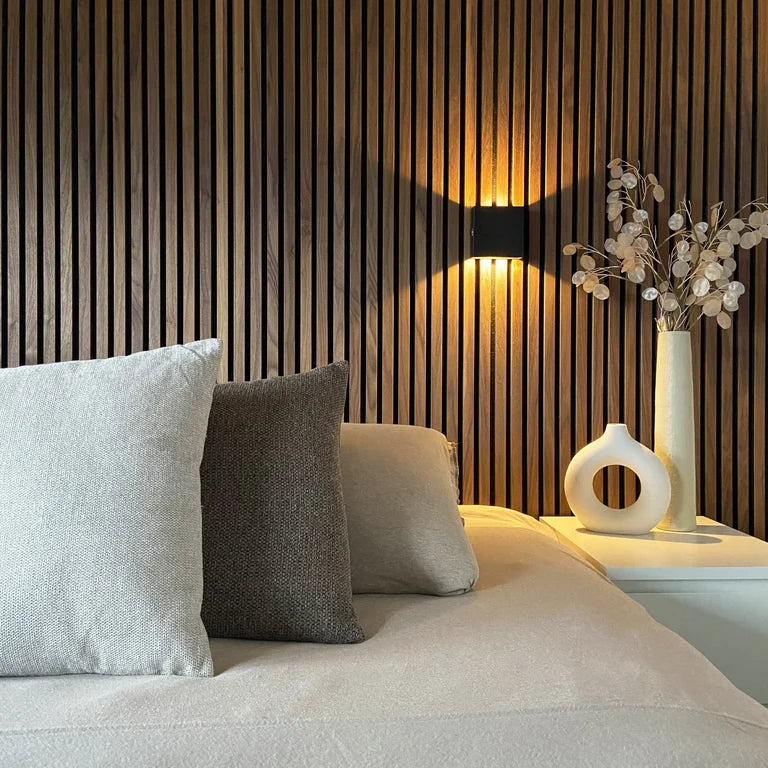 LUNIFY Boca compact aluminium wall light mounted on a slatted wood wall, casting a warm 2700K up-and-down glow. Wireless rechargeable LED wall lamp with motion sensor and magnetic mounting for wire-free indoor installation.