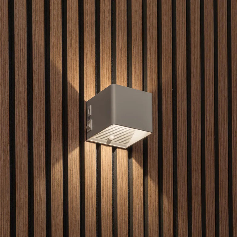 LUNIFY Boca compact aluminium wall light mounted on a slatted wood wall, casting a warm 2700K up-and-down glow. Wireless rechargeable LED wall lamp with motion sensor and magnetic mounting for wire-free indoor installation.