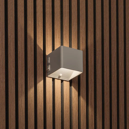 LUNIFY Boca compact aluminium wall light mounted on a slatted wood wall, casting a warm 2700K up-and-down glow. Wireless rechargeable LED wall lamp with motion sensor and magnetic mounting for wire-free indoor installation.