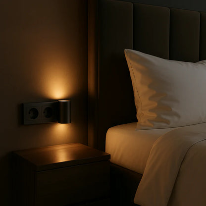PLUGIFY One plug-in wall light in matte finish mounted in a bedroom wall socket, casting warm ambient light onto a wooden bedside table with a remote control and ceramic mug beside it.