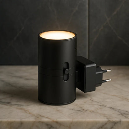 PLUGIFY One plug-in wall light in matte finish mounted in a bedroom wall socket, casting warm ambient light onto a wooden bedside table with a remote control and ceramic mug beside it.
