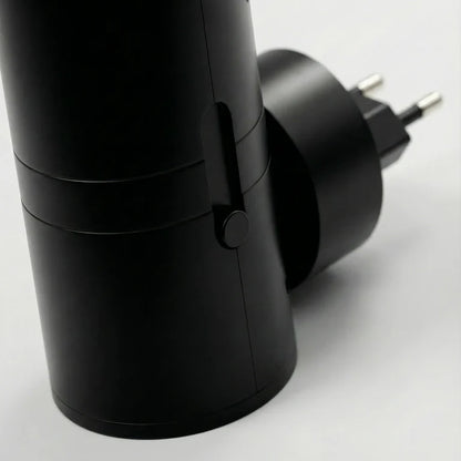 PLUGIFY One plug-in wall light in matte finish mounted in a bedroom wall socket, casting warm ambient light onto a wooden bedside table with a remote control and ceramic mug beside it.