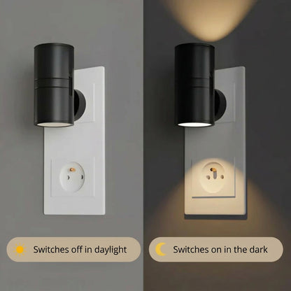 PLUGIFY One plug-in wall light in matte finish mounted in a bedroom wall socket, casting warm ambient light onto a wooden bedside table with a remote control and ceramic mug beside it.