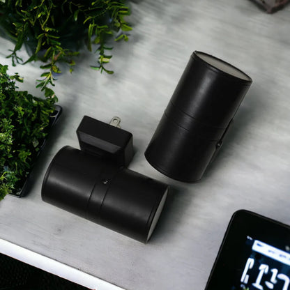 PLUGIFY One plug-in wall light in matte finish mounted in a bedroom wall socket, casting warm ambient light onto a wooden bedside table with a remote control and ceramic mug beside it.